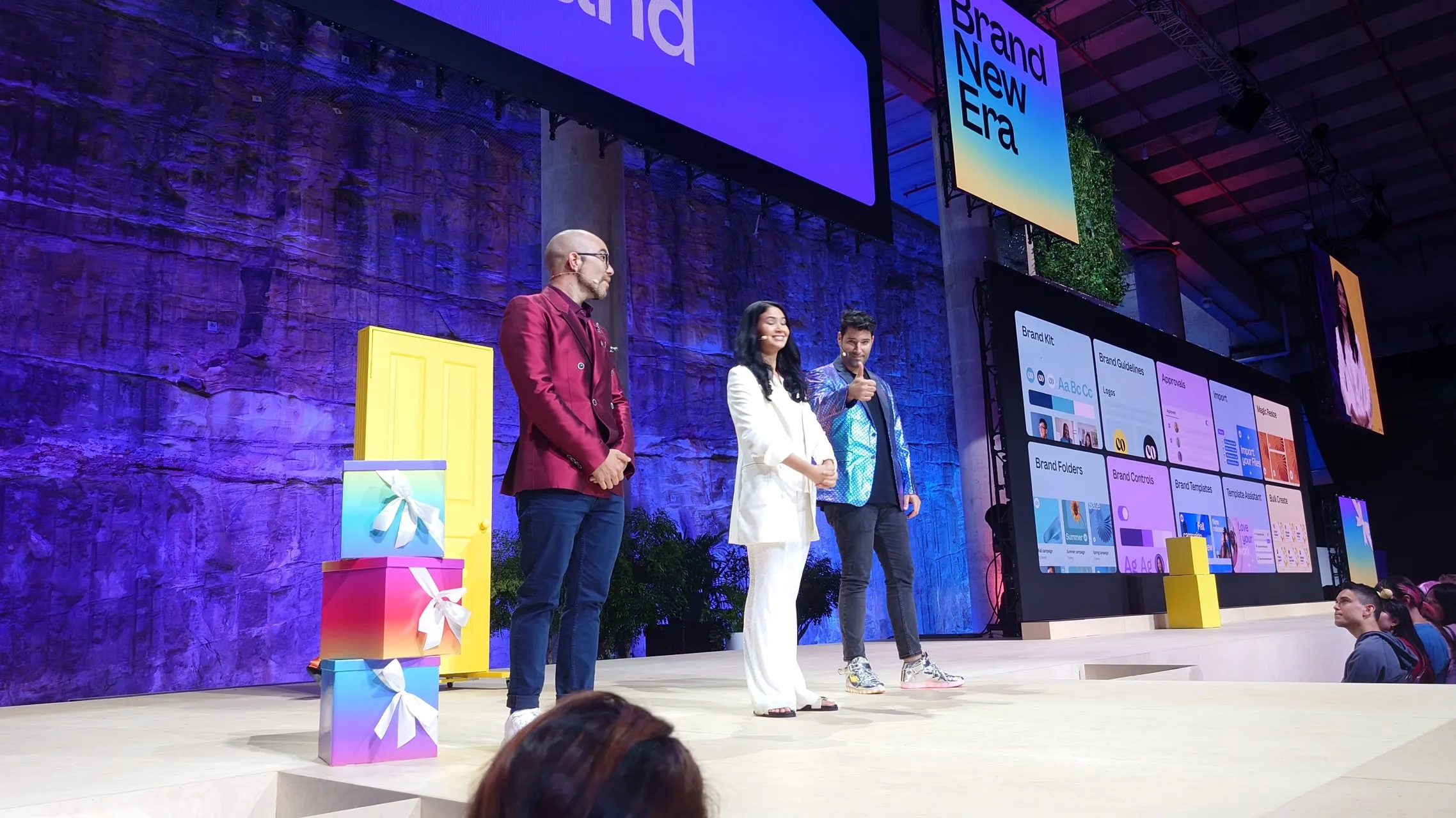Canva founders Mel (center), Cliff (right), and Cam (left) stand on stage with a large screen displaying the slogan "Brand New Era" in the background.